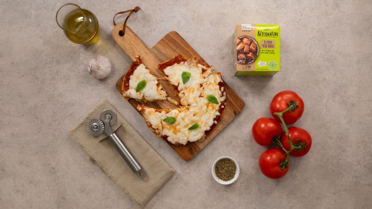 פיצה טופו מוכנה על קרש הגשה, לצד עגבניות, שמן זית, טופו ותבלינים.