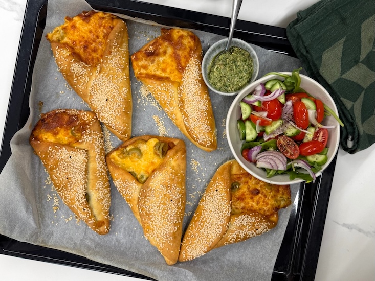 סמבוסק גבינה בקיפול קונוס מוכן על תבנית אפיה, לצד מטבל וסלט ירקות.