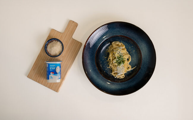 ספגטי ברוטב יוגורט בקערה, לצד קרש עם גבינת פרמזן וגביע יוגורט תנובה.